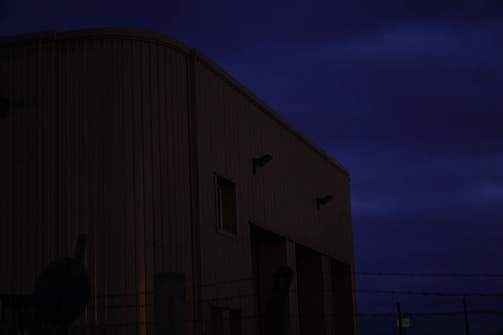 A large industrial building with a corrugated metal exterior is in semi-darkness. There is a barbed wire fence in the foreground, adding a sense of security or restriction. The sky is dark with heavy clouds, suggesting an evening or nighttime setting.