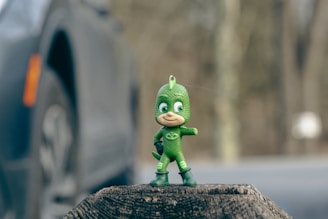 A small, green action figure standing on a tree stump. The figure is dressed in a green suit with a mask and boots and is striking a dynamic pose. In the background, there is a blurred image of a car and a forest setting.