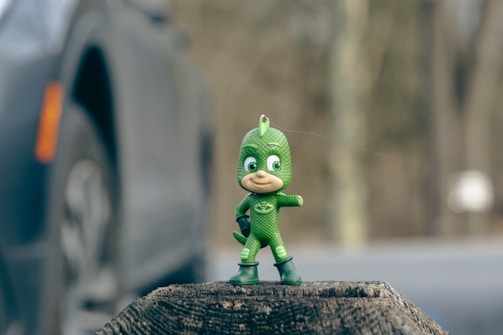 A small, green action figure standing on a tree stump. The figure is dressed in a green suit with a mask and boots and is striking a dynamic pose. In the background, there is a blurred image of a car and a forest setting.