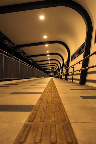 A long corridor with a tactile guiding path on the floor, illuminated by ceiling lights evenly spaced along the arc-shaped roof. The symmetrical design creates a sense of depth, while metallic railings line the sides.