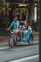 A man wearing a blue shirt and sunglasses rides a bicycle rickshaw on a city street, carrying a woman and a small child. The surroundings include trees and a building with a warmly lit interior, suggesting an urban, possibly commercial area.