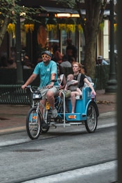 A man wearing a blue shirt and sunglasses rides a bicycle rickshaw on a city street, carrying a woman and a small child. The surroundings include trees and a building with a warmly lit interior, suggesting an urban, possibly commercial area.