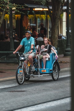 A man wearing a blue shirt and sunglasses rides a bicycle rickshaw on a city street, carrying a woman and a small child. The surroundings include trees and a building with a warmly lit interior, suggesting an urban, possibly commercial area.