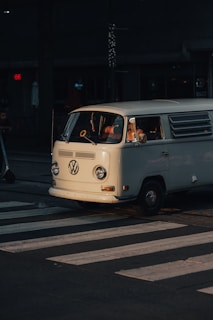 A classic white Volkswagen van is stopped at a pedestrian crossing, partially illuminated by warm sunlight. The van's vintage design is highlighted against the dark background of the street, with shadows enhancing its features. The setting suggests an urban environment, and there's a hint of a person inside the van.