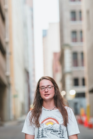 A young person with long hair and glasses stands in a city alleyway, wearing a graphic t-shirt featuring a colorful landscape design. The background displays blurred buildings with a mix of tan and gray tones and some out-of-focus street lights.