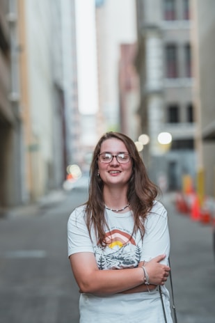 A person with long hair and glasses stands confidently with arms crossed in an urban alley. They wear a casual white t-shirt with a colorful graphic design. The background is blurred, with tall buildings lining the alley and some bright lights visible.