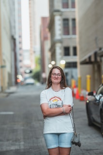 A young woman stands confidently in the middle of an empty urban street. She is wearing glasses, a white graphic T-shirt, and light denim shorts. Her arms are crossed, and she holds a small handbag in her left hand. The street is lined with tall buildings, with some visible construction cones and barriers to the right. The background is slightly blurred, highlighting the woman as the main focus.