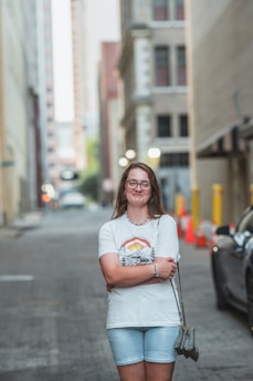 A young woman stands confidently in the middle of an empty urban street. She is wearing glasses, a white graphic T-shirt, and light denim shorts. Her arms are crossed, and she holds a small handbag in her left hand. The street is lined with tall buildings, with some visible construction cones and barriers to the right. The background is slightly blurred, highlighting the woman as the main focus.