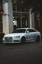 A sleek silver sedan is parked near an urban building with large windows. The car has a distinctive grille and black wheels, and the setting suggests an urban environment, possibly near a cafe or restaurant, as indicated by the interior lights visible through the windows.