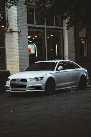 A sleek silver sedan is parked near an urban building with large windows. The car has a distinctive grille and black wheels, and the setting suggests an urban environment, possibly near a cafe or restaurant, as indicated by the interior lights visible through the windows.