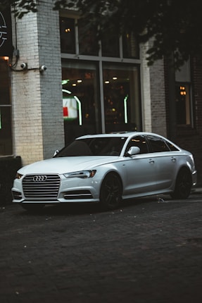 A sleek silver sedan is parked near an urban building with large windows. The car has a distinctive grille and black wheels, and the setting suggests an urban environment, possibly near a cafe or restaurant, as indicated by the interior lights visible through the windows.