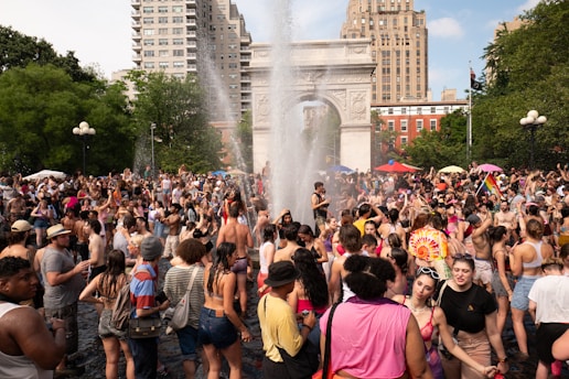 A large crowd of people gathers near a prominent stone arch in a park setting, surrounded by trees and buildings. Many in the crowd appear to be celebrating, with some holding rainbow flags and wearing colorful outfits, suggesting a festival or parade atmosphere. The central fountain sprays water into the air, and people are enjoying themselves in the warm, sunny weather.
