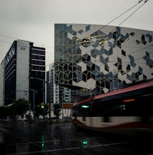 An urban scene featuring a modern building with a geometric facade pattern, alongside a streetcar moving along wet tracks with reflections on the pavement. High-rise buildings surround the area, and traffic lights are visible in the dim lighting, suggesting a gloomy, overcast day.