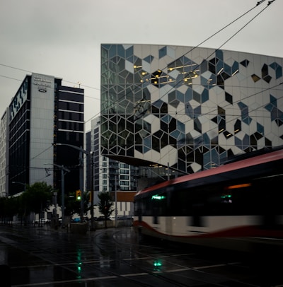 An urban scene featuring a modern building with a geometric facade pattern, alongside a streetcar moving along wet tracks with reflections on the pavement. High-rise buildings surround the area, and traffic lights are visible in the dim lighting, suggesting a gloomy, overcast day.