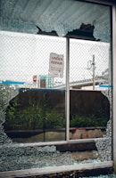 A broken glass window with shattered fragments surrounds a sign that reads 'Under Repair, Thank You For Your Patience'. Behind the window, there is a mesh fence, some grass, and an urban setting with signs and structures visible in the background.