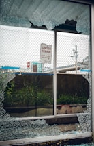 A broken glass window with shattered fragments surrounds a sign that reads 'Under Repair, Thank You For Your Patience'. Behind the window, there is a mesh fence, some grass, and an urban setting with signs and structures visible in the background.