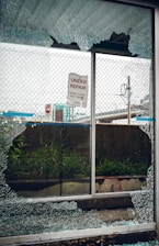 A broken glass window with shattered fragments surrounds a sign that reads 'Under Repair, Thank You For Your Patience'. Behind the window, there is a mesh fence, some grass, and an urban setting with signs and structures visible in the background.