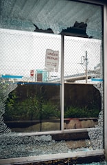 A broken glass window with shattered fragments surrounds a sign that reads 'Under Repair, Thank You For Your Patience'. Behind the window, there is a mesh fence, some grass, and an urban setting with signs and structures visible in the background.