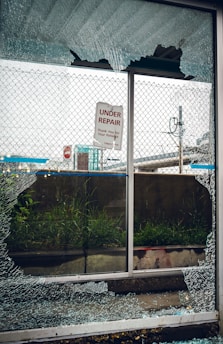 A broken glass window with shattered fragments surrounds a sign that reads 'Under Repair, Thank You For Your Patience'. Behind the window, there is a mesh fence, some grass, and an urban setting with signs and structures visible in the background.