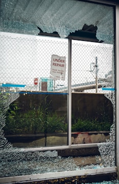 A broken glass window with shattered fragments surrounds a sign that reads 'Under Repair, Thank You For Your Patience'. Behind the window, there is a mesh fence, some grass, and an urban setting with signs and structures visible in the background.