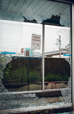 A broken glass window with shattered fragments surrounds a sign that reads 'Under Repair, Thank You For Your Patience'. Behind the window, there is a mesh fence, some grass, and an urban setting with signs and structures visible in the background.