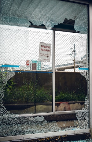 A broken glass window with shattered fragments surrounds a sign that reads 'Under Repair, Thank You For Your Patience'. Behind the window, there is a mesh fence, some grass, and an urban setting with signs and structures visible in the background.