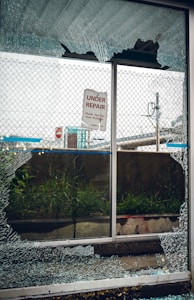 A broken glass window with shattered fragments surrounds a sign that reads 'Under Repair, Thank You For Your Patience'. Behind the window, there is a mesh fence, some grass, and an urban setting with signs and structures visible in the background.