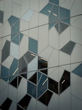 An abstract architectural facade featuring a series of geometric patterns created by irregularly shaped glass panels bordered by metal frames. The panels have varying shades and reflections, adding to the complexity of the design.