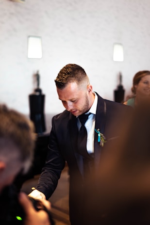 A man in a formal dark suit looks downward with a thoughtful expression. A boutonniere decorates his suit, and ambient light casts soft shadows on the neutral-colored wall behind. Another person is slightly visible in the blurry foreground, adding a candid feel to the scene.