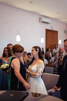A bride in a lace wedding dress is interacting warmly with a guest in a dark dress holding a small bouquet. The room is filled with other seated guests, some of whom are watching the interaction while others are occupied with their phones.