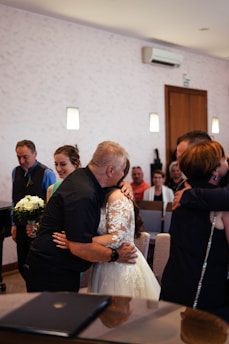 A group of people in a room, with some in formal attire. Two people are embracing in the center, one of whom is wearing a lace white dress. A small bouquet of flowers is visible in the hands of a person on the left. There is a piano in the foreground, and the background shows a light-colored wall with sconces and seated guests.