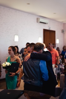 A group of people gathered indoors, where two individuals appear to be in an emotional embrace in the foreground. Several other people are in the vicinity, some holding flowers, suggesting a formal or celebratory event. The room is softly lit with a minimal and clean design.