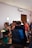 A group of people gathered indoors, where two individuals appear to be in an emotional embrace in the foreground. Several other people are in the vicinity, some holding flowers, suggesting a formal or celebratory event. The room is softly lit with a minimal and clean design.