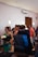 A group of people gathered indoors, where two individuals appear to be in an emotional embrace in the foreground. Several other people are in the vicinity, some holding flowers, suggesting a formal or celebratory event. The room is softly lit with a minimal and clean design.