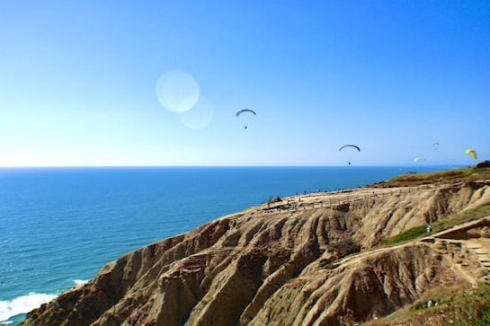 A picturesque coastal scene with a clear blue sky and several colorful paragliders soaring over the ocean and rocky cliffs. The rugged terrain contrasts with the vast expanse of the sea, creating a dynamic and adventurous atmosphere.