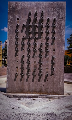 A concrete wall with multiple protruding metal nozzles arranged in a symmetrical pattern. The structure casts distinct shadows on the wall, and there are patches of discoloration and weathering. The base of the wall includes a drainage area, indicating possible use as a fountain. The environment around includes paved walkways and distant trees under a bright blue sky.