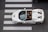 Aerial view of a white convertible sports car with two passengers driving over a pedestrian crossing on a city street. The car's sleek design and open roof are clearly visible, and the passengers are seated comfortably, one wearing a striped shirt.