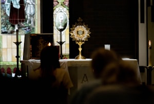 A golden monstrance is prominently placed on an altar with a white cloth, flanked by lit candles. In the background, there are stained glass windows depicting religious scenes. Silhouettes of people appear in the foreground, suggesting a reverent or prayerful atmosphere.
