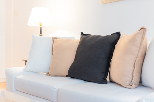 A cozy living room setup featuring a white sofa adorned with several pillows in beige, black, and cream colors. A warm-lit lamp is situated on the left side, adding a soft glow to the space.