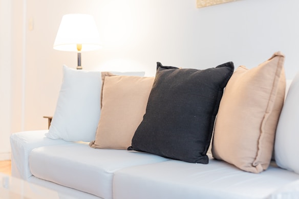 A cozy living room setup featuring a white sofa adorned with several pillows in beige, black, and cream colors. A warm-lit lamp is situated on the left side, adding a soft glow to the space.