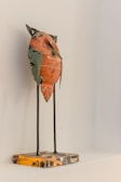 A small sculpture of a stylized bird made from recycled materials, including a recognizable painted orange metal piece with text. The bird stands on thin metal legs attached to a square, patterned base. The wall behind the sculpture is a neutral light color.