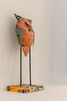A small sculpture of a stylized bird made from recycled materials, including a recognizable painted orange metal piece with text. The bird stands on thin metal legs attached to a square, patterned base. The wall behind the sculpture is a neutral light color.
