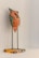 A small sculpture of a stylized bird made from recycled materials, including a recognizable painted orange metal piece with text. The bird stands on thin metal legs attached to a square, patterned base. The wall behind the sculpture is a neutral light color.