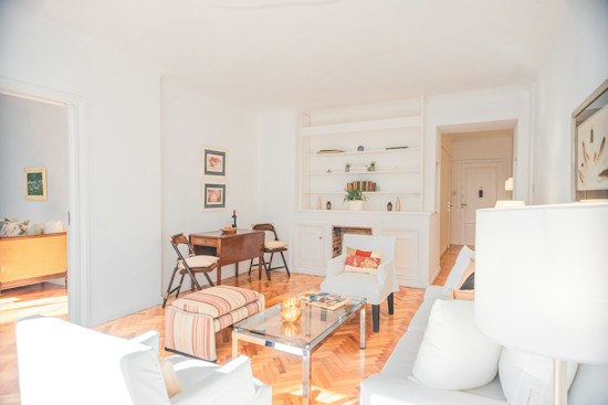A bright and airy living room features light-colored walls and wooden flooring. The space is furnished with white armchairs, a glass coffee table, and a striped ottoman. A wooden desk with chairs is placed near the wall, adorned with framed art. Built-in shelves above a fireplace display decorative items and plants. Natural light floods the room from the adjacent area.