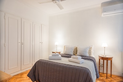 A neatly made bed is centered in a bright room with a white duvet, several pillows, and towels placed on top. The room features a simple decor with two bedside tables, each holding a lamp. A ceiling fan and an air conditioning unit are visible. The floor is made of hardwood, and there are built-in wardrobes on one wall.