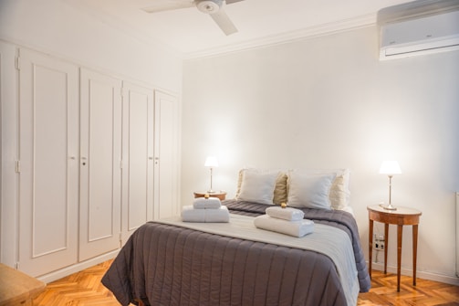 A neatly made bed is centered in a bright room with a white duvet, several pillows, and towels placed on top. The room features a simple decor with two bedside tables, each holding a lamp. A ceiling fan and an air conditioning unit are visible. The floor is made of hardwood, and there are built-in wardrobes on one wall.