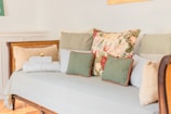 A wooden daybed adorned with a variety of cushions, including a floral-patterned pillow, several green and beige pillows, and neatly folded white towels. The setting features a soft, pastel color palette, creating a cozy and inviting atmosphere.