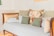 A wooden daybed adorned with a variety of cushions, including a floral-patterned pillow, several green and beige pillows, and neatly folded white towels. The setting features a soft, pastel color palette, creating a cozy and inviting atmosphere.