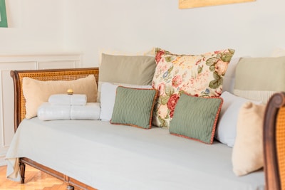 A wooden daybed adorned with a variety of cushions, including a floral-patterned pillow, several green and beige pillows, and neatly folded white towels. The setting features a soft, pastel color palette, creating a cozy and inviting atmosphere.