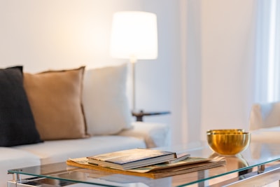 A cozy living room setting with a sofa adorned with a black and a beige cushion. On a sleek glass coffee table in the foreground, a stack of magazines is neatly placed next to a shiny brass or gold-colored bowl. A soft, warm ambiance is created by a lit lamp in the background.